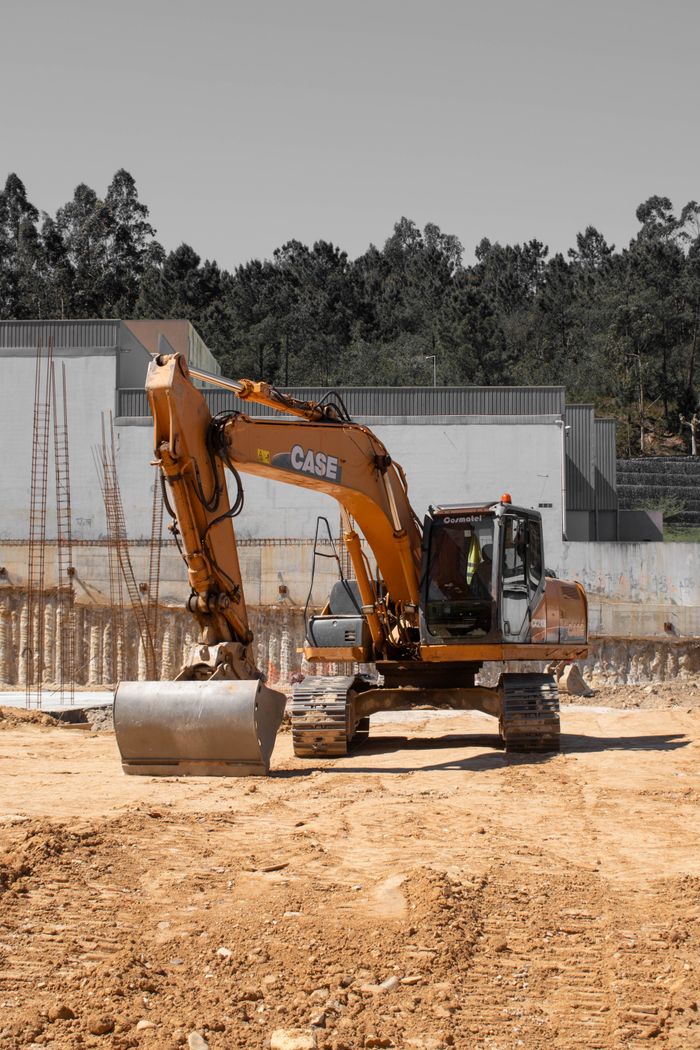 An excavator at a construction site against a forested backdrop under a clear sky.