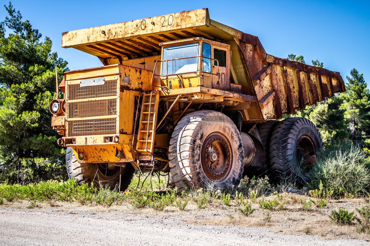 Dirty yellow tipper truck parked on dusty rural road near green trees against cloudless blue sky