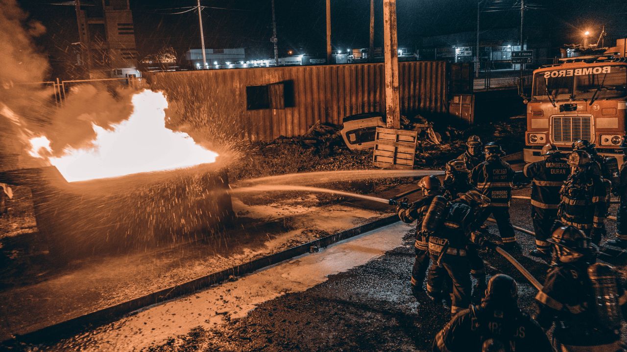 Firefighters combat a raging fire at night using hoses in an industrial urban area.
